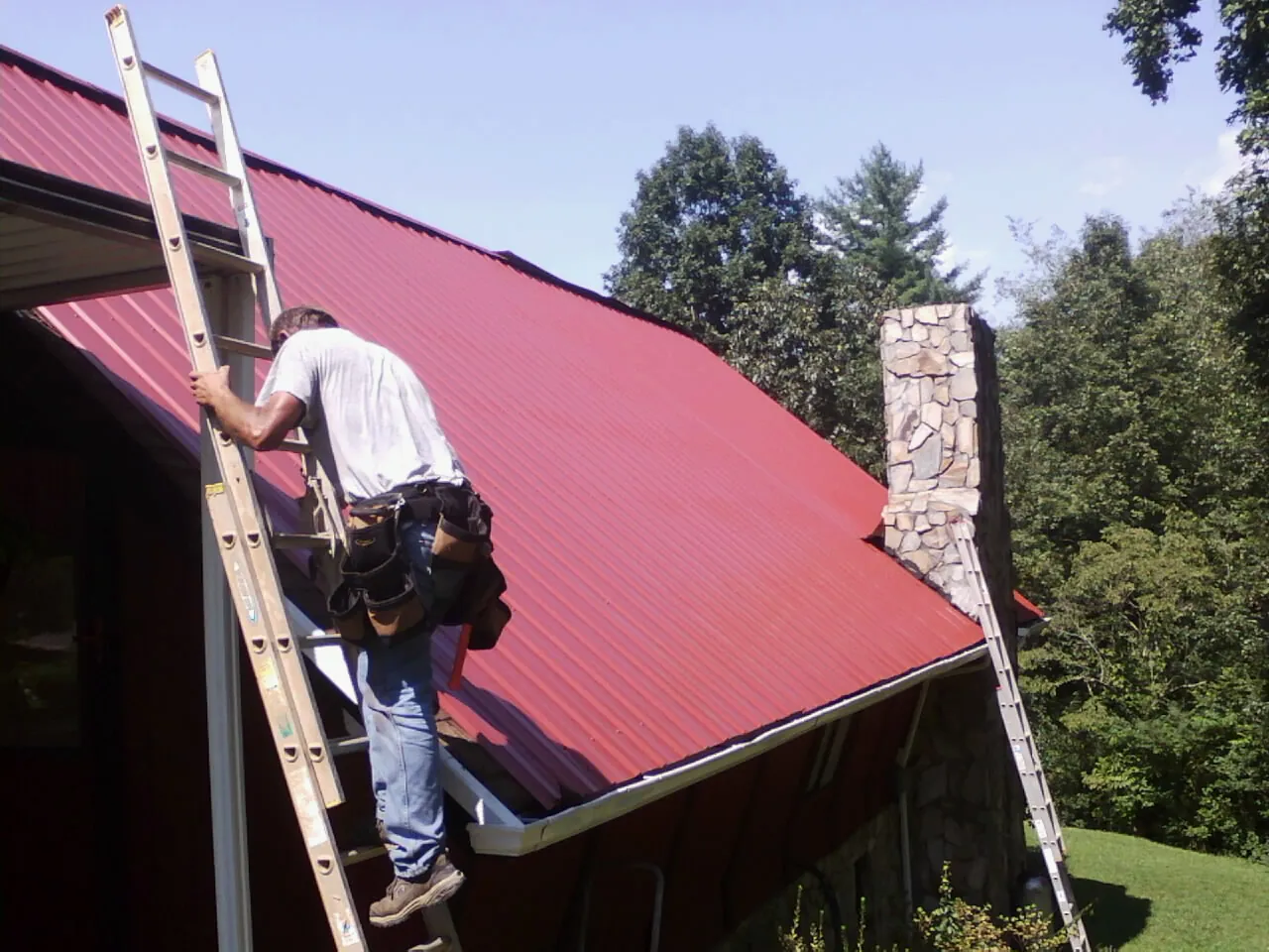 Roofer on a ladder beside a red metal roof and stone chimney — Soffit & Fascia Repair by Roofing Brothers in Starrucca